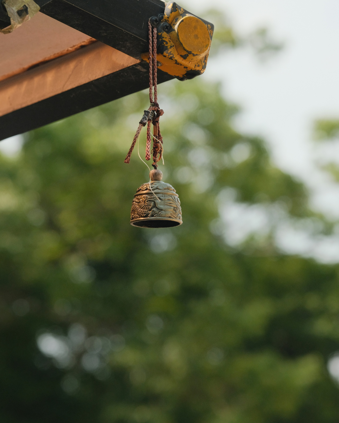 A beautiful golden bell hanging from a roof. The background is green from foliage and trees. 